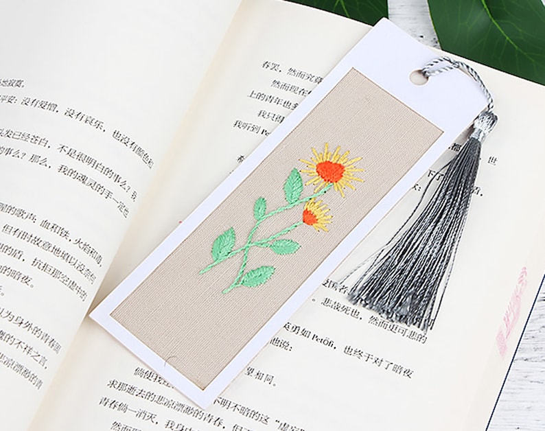 May include: A beige bookmark with a white border and a tassel. The bookmark features an embroidered design of a green stem with two yellow flowers.