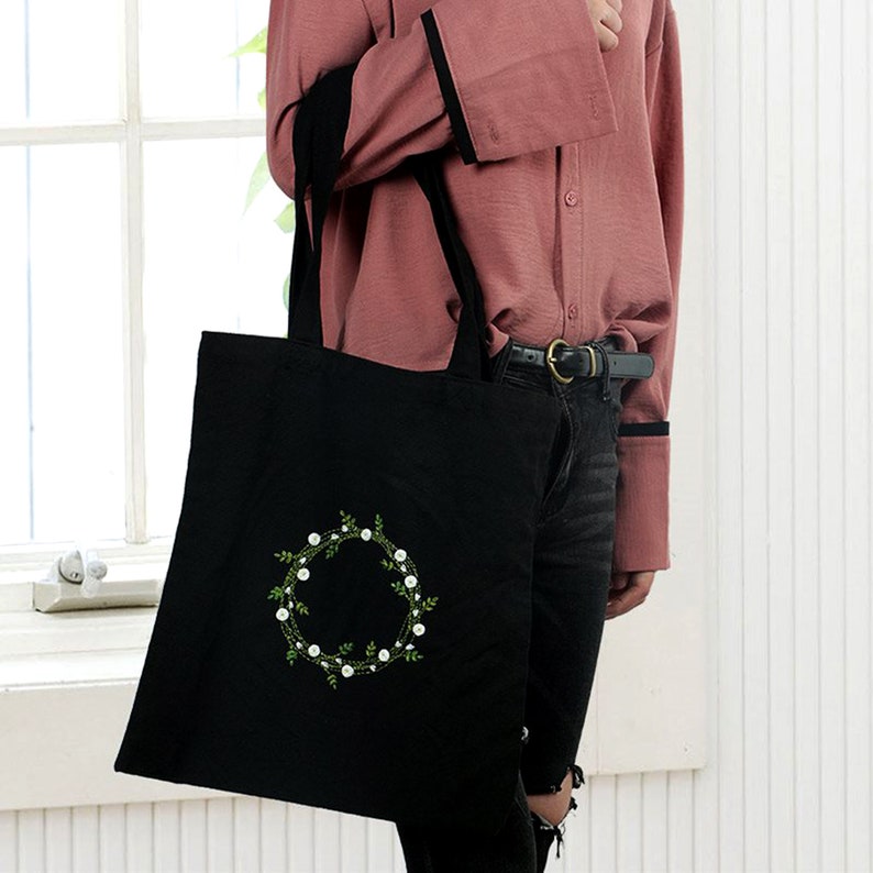 May include: A black canvas tote bag with a white floral wreath design. The wreath is embroidered with white flowers and green leaves.