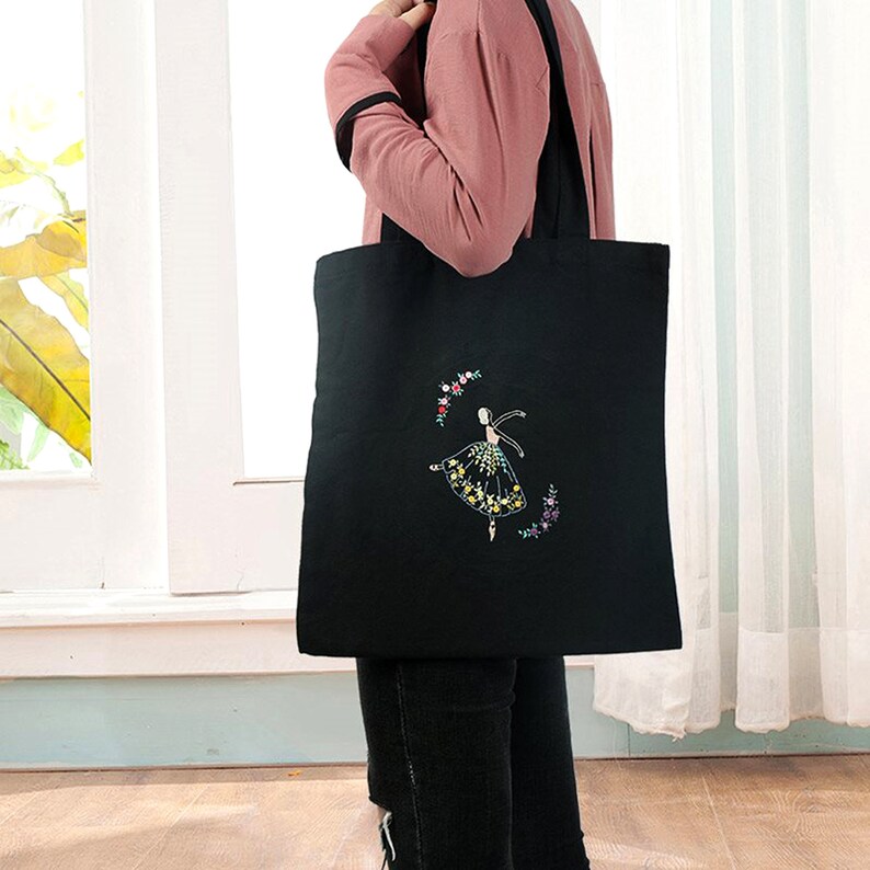 May include: Black canvas tote bag with an embroidered design of a ballerina in a floral skirt. The ballerina is surrounded by a wreath of flowers.