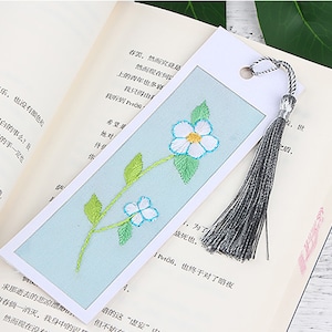 May include: A blue and white embroidered bookmark with a floral design and a grey tassel. The bookmark is on a book with text in a foreign language.