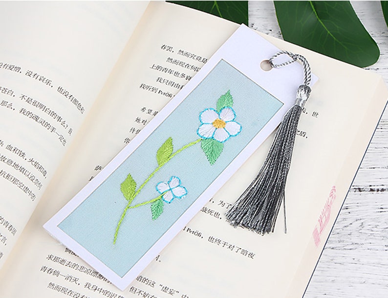 May include: A blue and white embroidered bookmark with a floral design and a grey tassel. The bookmark is on a book with text in a foreign language.