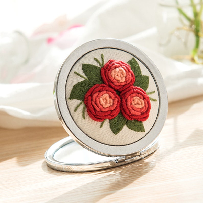 Op de afbeelding: Een kleine, ronde spiegel met een zilveren frame en een witte stoffen achtergrond geborduurd met drie rode en roze rozen en groene bladeren.