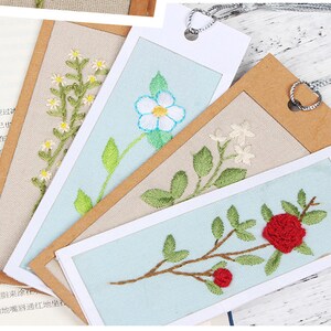 May include: Four embroidered bookmarks featuring different floral designs. The bookmarks are displayed on a light brown background. The designs include a white flower with a blue centre, a red rose, a white flower with a green stem, and a white flower with a green stem and leaves.
