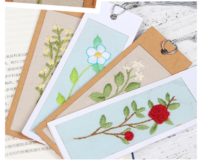 May include: Four embroidered bookmarks featuring different floral designs. The bookmarks are displayed on a light brown background. The designs include a white flower with a blue centre, a red rose, a white flower with a green stem, and a white flower with a green stem and leaves.