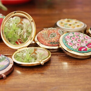 May include: A set of six round, gold-framed compact mirrors with embroidered floral designs. The mirrors are open, revealing a clear reflection of green plants.