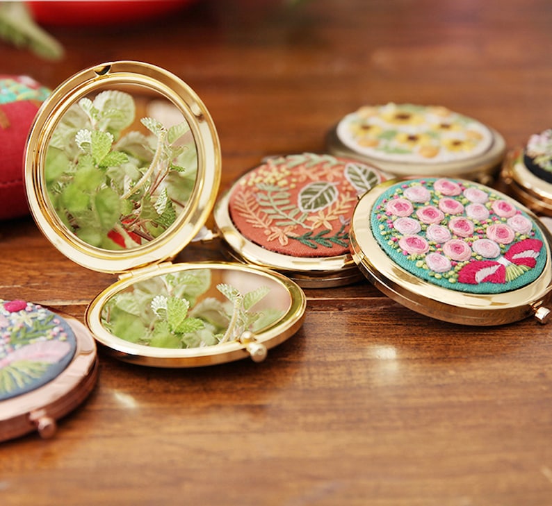 May include: A set of six round, gold-framed compact mirrors with embroidered floral designs. The mirrors are open, revealing a clear reflection of green plants.
