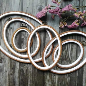 Peut inclure: Plusieurs cercles à broder ovales de différentes tailles. Ils présentent un cadre en bois brun clair avec un intérieur blanc et des fixations en laiton vieilli. L'ensemble est disposé sur un fond en bois rustique, avec des fleurs violettes séchées.