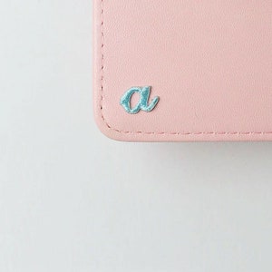 May include: A pink leather wallet with a light blue embroidered letter "a" on the front.