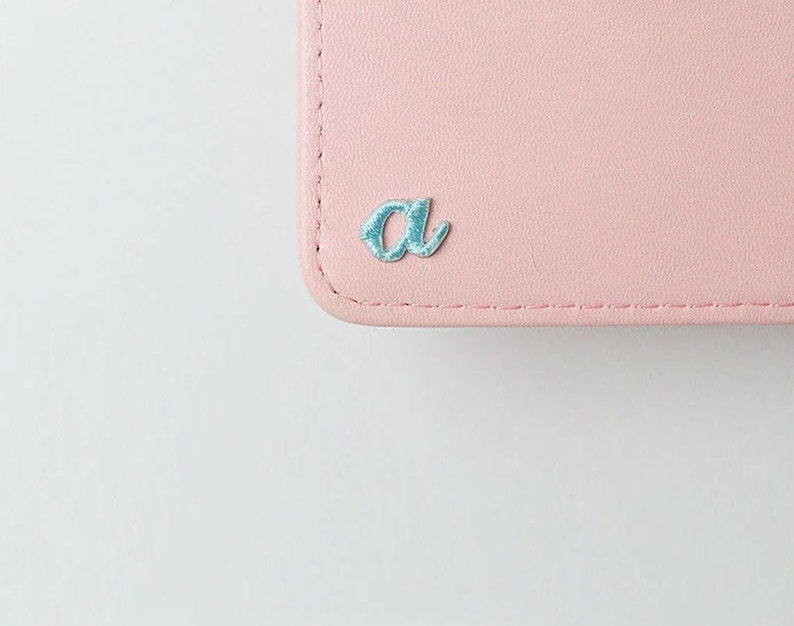 May include: A pink leather wallet with a light blue embroidered letter "a" on the front.