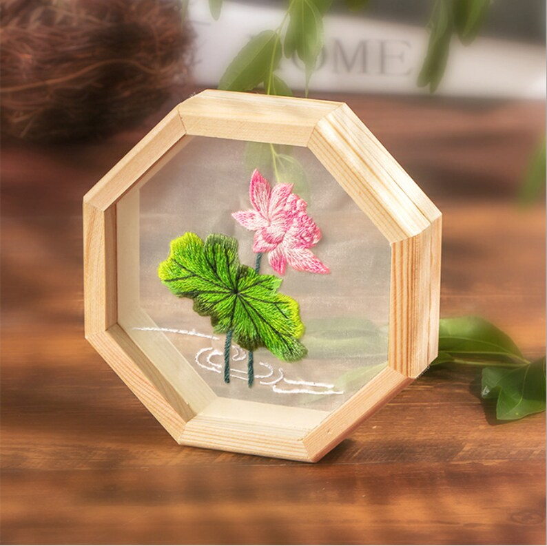 Peut inclure: Une broderie encadr&eacute;e repr&eacute;sentant un lotus rose avec des feuilles vertes, sur fond blanc. Le cadre est en bois clair et a une forme octogonale.