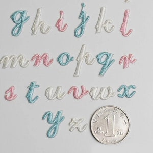 May include: A set of embroidered letters in cursive script, with each letter in a different color: light blue, pink, and white. The letters are arranged in alphabetical order, with the letter 'g' at the top left and the letter 'x' at the bottom right. A Chinese coin is also visible in the image.