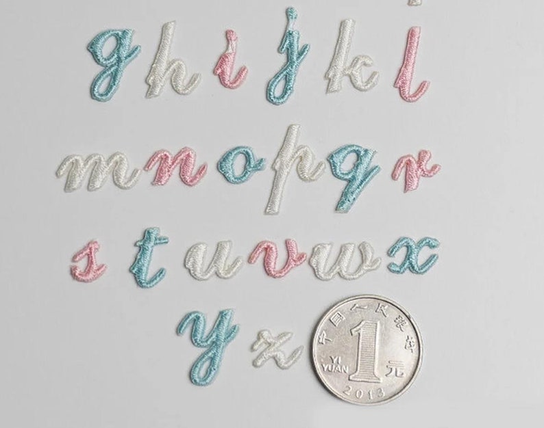 May include: A set of embroidered letters in cursive script, with each letter in a different color: light blue, pink, and white. The letters are arranged in alphabetical order, with the letter 'g' at the top left and the letter 'x' at the bottom right. A Chinese coin is also visible in the image.
