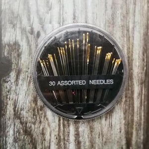 May include: A round plastic container with a black lid and a clear bottom. The container holds 30 assorted needles. The container has the text "30 Assorted Needles" printed on the front.