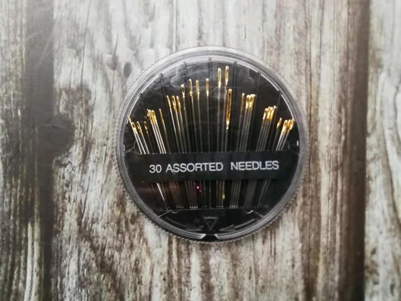 May include: A round plastic container with a black lid and a clear bottom. The container holds 30 assorted needles. The container has the text "30 Assorted Needles" printed on the front.
