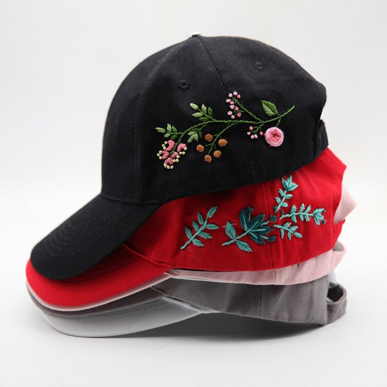 May include: A stack of four baseball caps with embroidered floral designs. The top cap is black with a green and pink floral design. The second cap is red with a green floral design. The third cap is pink and the bottom cap is gray.