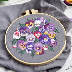 May include: A close-up of a hand-stitched embroidery hoop with a floral design. The hoop is made of wood and has a grey fabric background. The embroidery features a bouquet of purple, white, and yellow pansies.