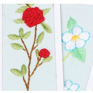 May include: Two embroidered fabric panels featuring floral designs. The first panel depicts a red rose with green leaves and stems. The second panel shows a white flower with a yellow centre and green leaves.