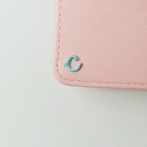 May include: A pale pink leather wallet with a light blue embroidered letter "C" on the front.