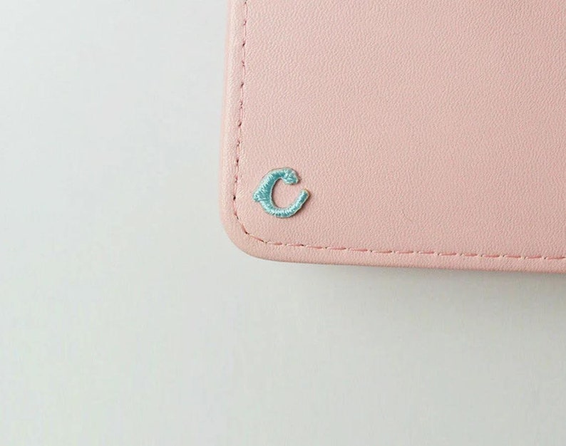 May include: A pale pink leather wallet with a light blue embroidered letter "C" on the front.