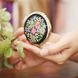May include: A round, gold-coloured compact mirror with a black background and a colourful floral embroidery design.
