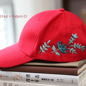 May include: Red baseball cap with a green floral embroidery design. The cap is sitting on a stack of books.