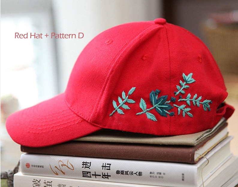 May include: Red baseball cap with a green floral embroidery design. The cap is sitting on a stack of books.