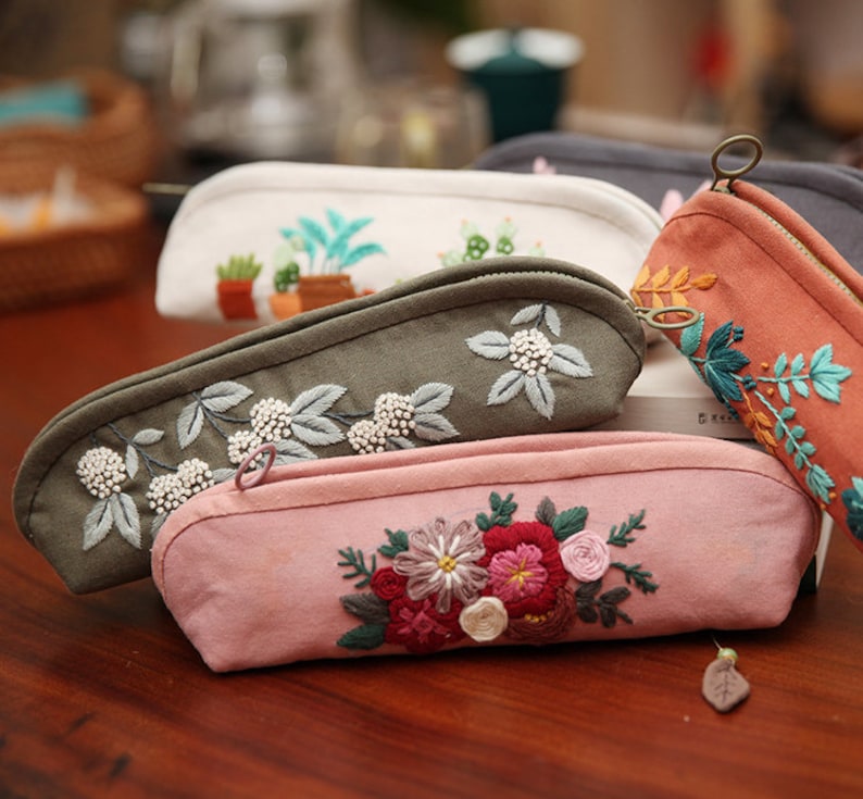 May include: Five embroidered pencil cases with different floral designs. The cases are made of a soft, canvas-like material and have a zipper closure. The designs include a pink floral pattern, a green floral pattern with white beads, a white floral pattern with green plants, a gray floral pattern, and an orange floral pattern.