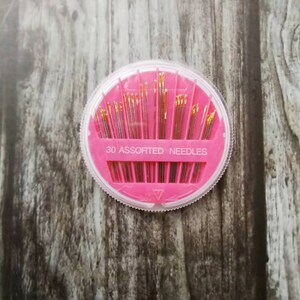 May include: A pink plastic container with a clear lid holds 30 assorted needles. The container has the text "30 Assorted Needles" printed on it.