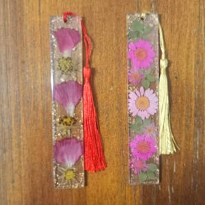 May include: Two rectangular resin bookmarks featuring embedded pressed flowers. The left bookmark contains pink petals and yellow centers with a red tassel. The right bookmark displays pink and purple daisy-like flowers with green leaves and a gold tassel.