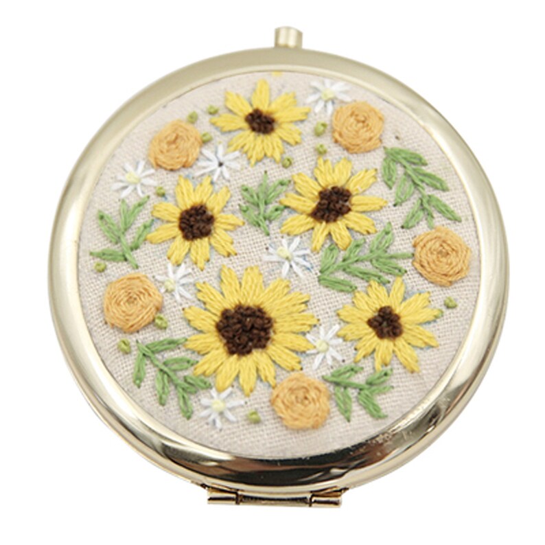 May include: A round, gold-framed mirror with a white fabric background embroidered with yellow sunflowers, green leaves, and white and yellow flowers.