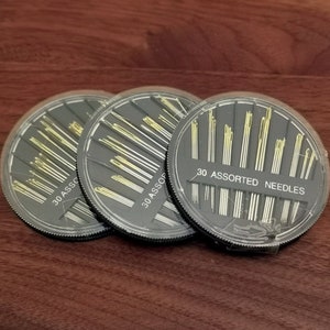May include: Three round black plastic containers filled with assorted needles. Each container has a label that reads "30 Assorted Needles".