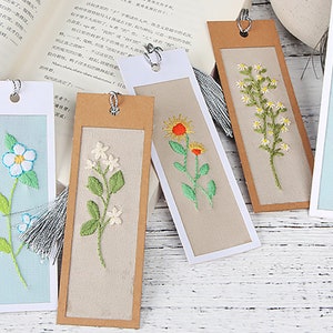 May include: Five handmade bookmarks featuring various floral embroidery designs. The bookmarks are crafted from brown and white cardstock and each has a tassel at the top.