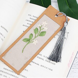 May include: A brown bookmark with a white fabric insert featuring an embroidered floral design. The bookmark has a grey tassel.