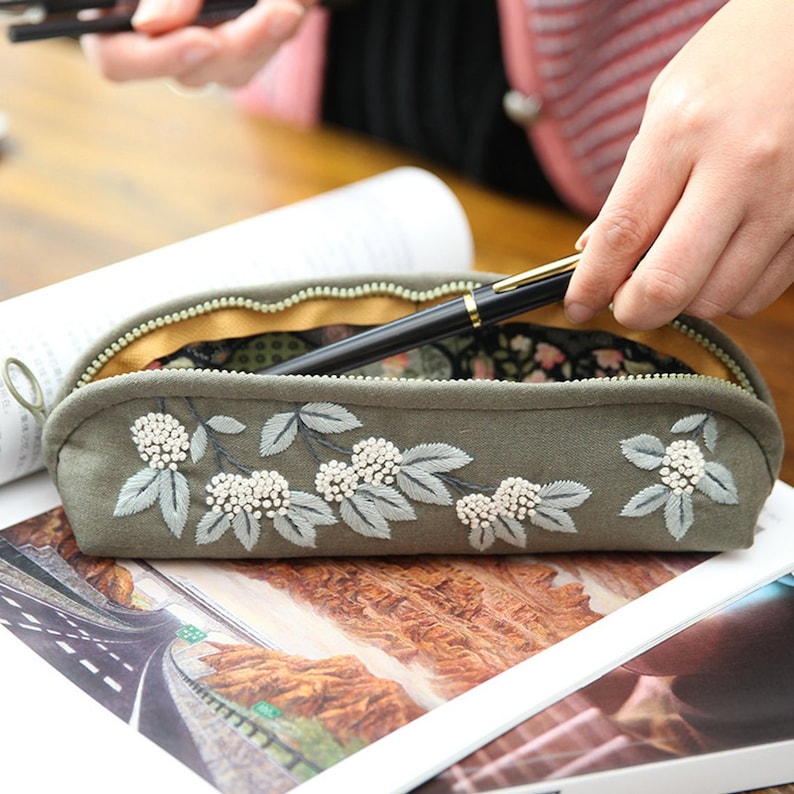 May include: A green fabric pencil case with a zipper closure and embroidered white flowers with green leaves. The case is open and a black pen is inside.