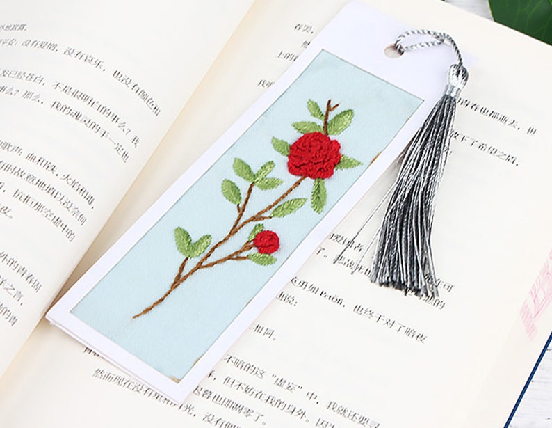 May include: A blue bookmark with a white border and a red embroidered flower design. The bookmark has a silver tassel.