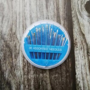 May include: A round, blue plastic container with a clear lid holds 30 assorted needles. The container has "30 ASSORTED NEEDLES" printed on it.