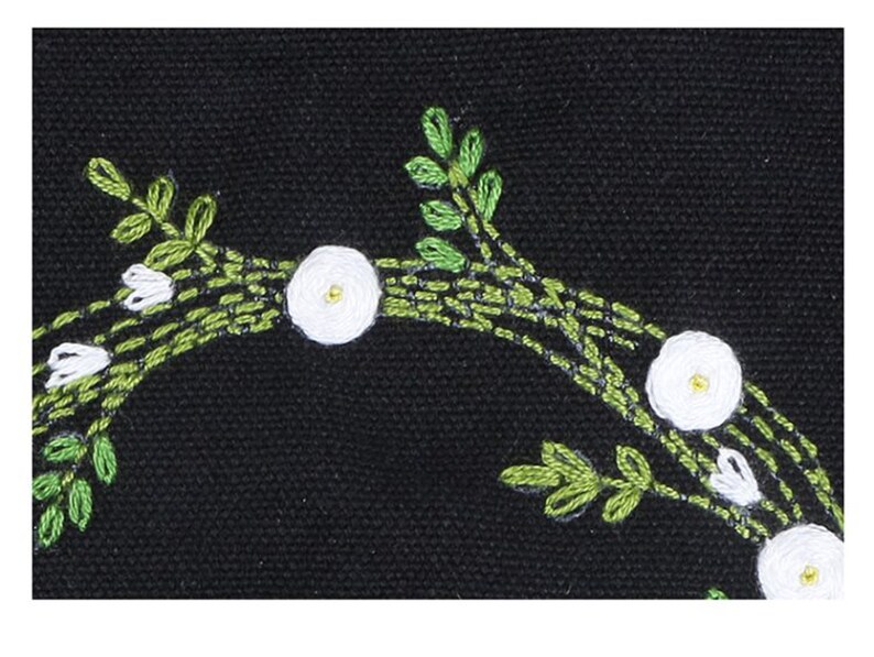 May include: Close-up of a black fabric with white flowers and green leaves embroidered on it. The flowers are simple and have a yellow center.
