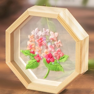 Könnte beinhalten: Eine gerahmte Stickerei mit rosa und grünen Blumen und schimmernden Pailletten. Der Rahmen ist aus hellem Holz und hat eine achteckige Form.
