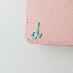 May include: A light pink leather wallet with a light blue embroidered letter "d" on the front.