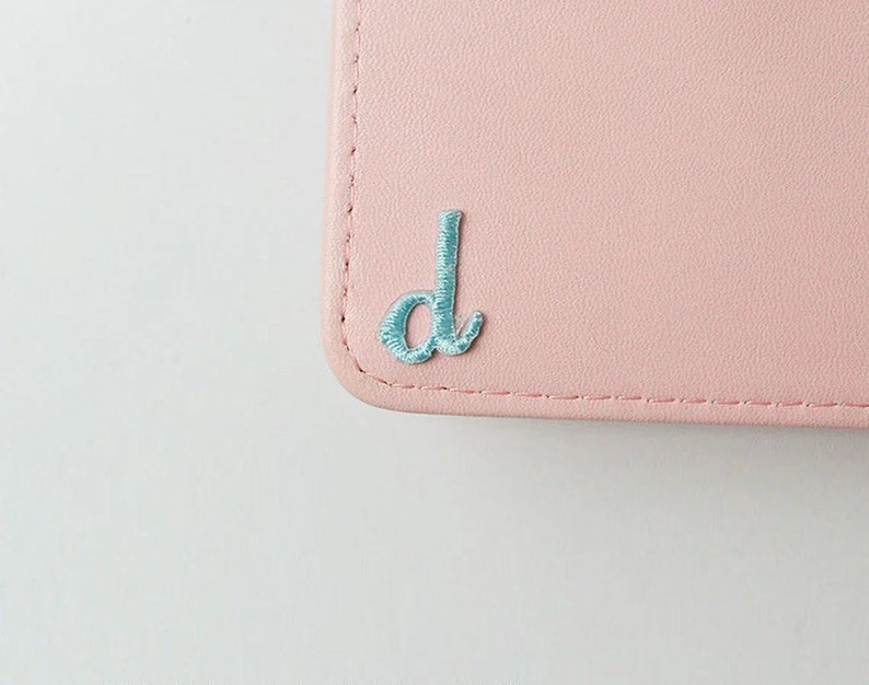 May include: A light pink leather wallet with a light blue embroidered letter "d" on the front.