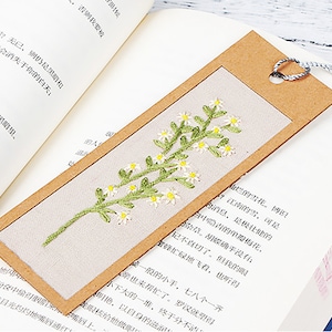 May include: A brown bookmark with a white fabric insert featuring an embroidered floral design of white flowers and green leaves. The bookmark has a brown paper border and a silver tassel.