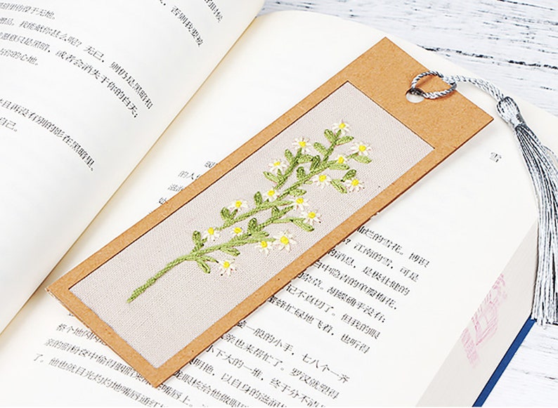 May include: A brown bookmark with a white fabric insert featuring an embroidered floral design of white flowers and green leaves. The bookmark has a brown paper border and a silver tassel.