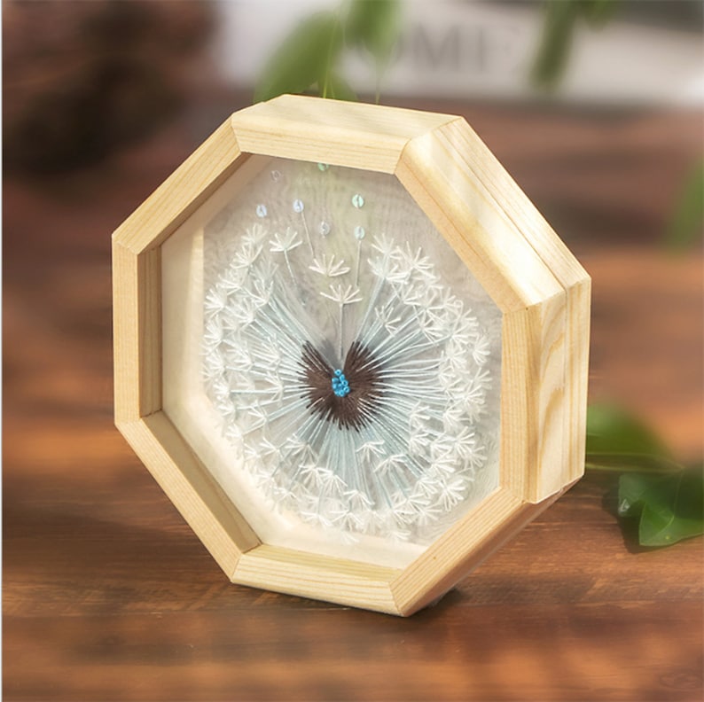 Peut inclure: Une fleur de pissenlit blanche avec un papillon bleu et brun au centre, encadr&eacute;e dans un octogone en bois clair.