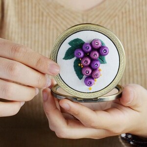 Op de afbeelding: Een kleine, ronde, goudkleurige compactspiegel met een wit interieur. Het interieur heeft een handgestikte borduursel van een tros paarse druiven met groene bladeren.