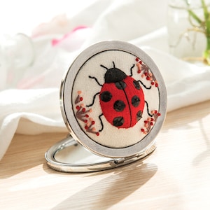 Op de afbeelding: Een zilveren compact spiegel met een witte stoffen hoes met een geborduurde rode lieveheersbeestje met zwarte stippen en roze bloemen.
