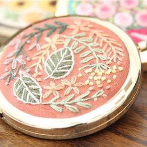 May include: A round, gold-framed compact mirror with a pink fabric interior. The fabric is embroidered with a floral design featuring green leaves, pink and yellow flowers, and white accents.