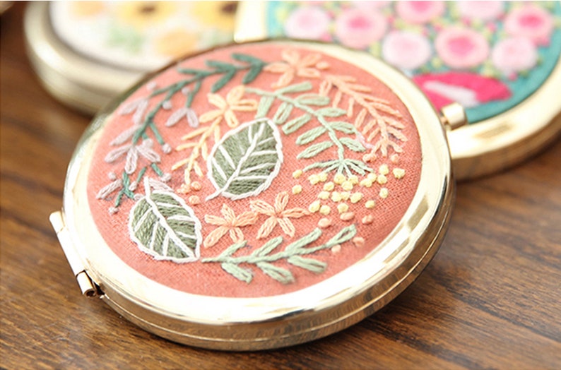 May include: A round, gold-framed compact mirror with a pink fabric interior. The fabric is embroidered with a floral design featuring green leaves, pink and yellow flowers, and white accents.