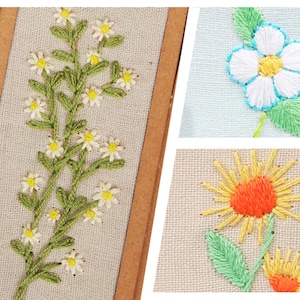 May include: Three embroidered floral designs on a beige fabric background. The first design is a cluster of white daisies with green stems and leaves. The second design is a single white flower with a yellow centre and blue outline. The third design is a yellow sunflower with an orange centre and green leaves.