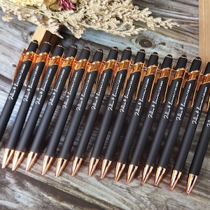 Op de afbeelding: Een groep van 16 zwarte pennen met roségouden accenten en een witte inscriptie die "Palm & Lauren" op elke pen staat.
