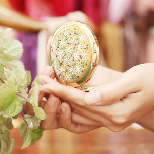 May include: A gold compact mirror with a floral design. The mirror is decorated with white daisies and purple berries on a yellow background.